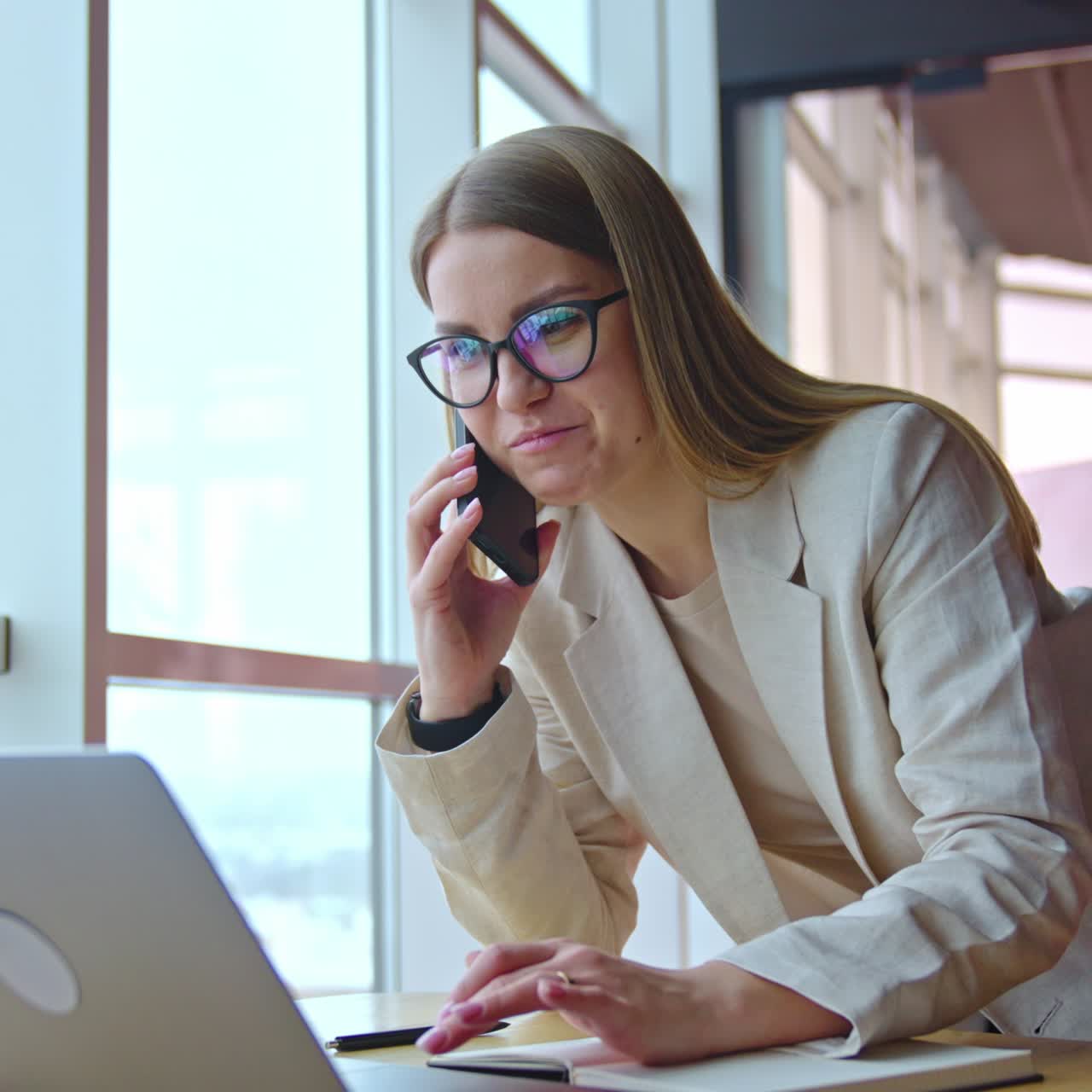Lady in light suit standing leaned over the desk. Smiling woman searches something on laptop and speaks on the phone