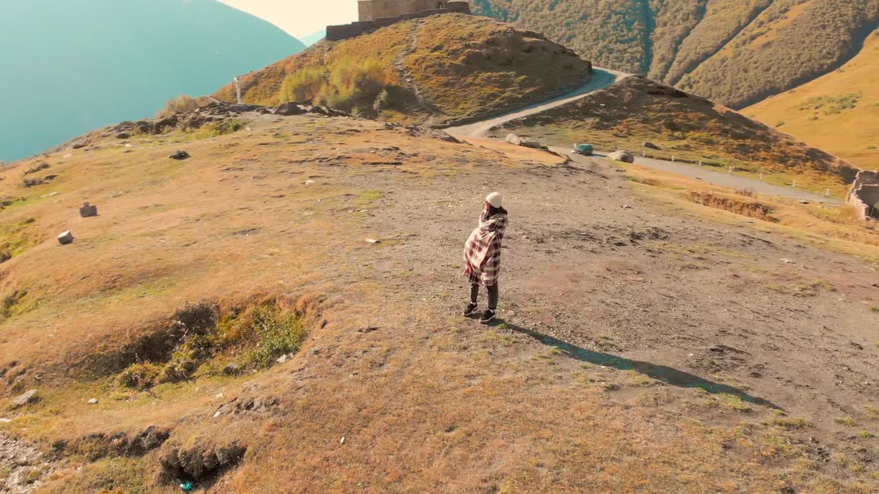 una mujer caucásica está emocionada en las montañas con la iglesia de la trinidad de kazbegi en el fondo.
