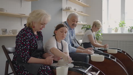 Medium-shot-of-middle-aged-ceramic-artist-teaching-group-elderly-Caucasian-woman-and-senior-man-how-to-wedge-clay-sitting-at-desk-in-art-studio.-People-enjoying-talking-at-work