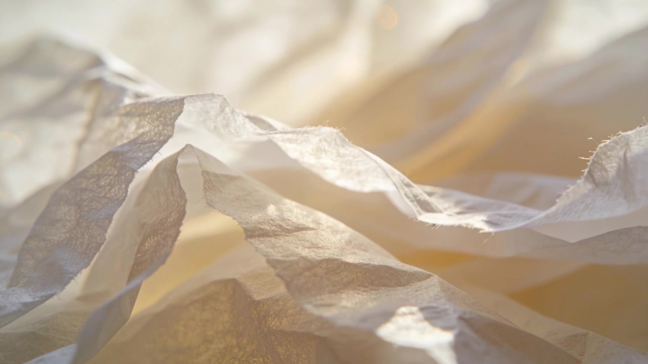 Reacting to warm backlight shift, camera revealing crumpled thin paper folds on studio table, lamp