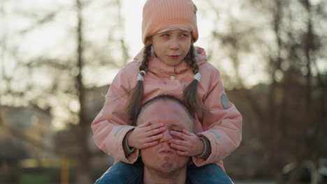 serene moment as a little girl in a pink cap calmly covers her father's eyes while sitting on his shoulders in a park