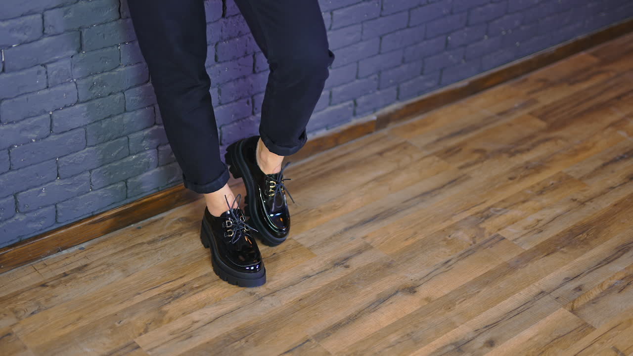 Woman wearing black massive shoes with laces and tractor soles. Lady stands leaned on the blue wall demonstrating fashionable footwear.