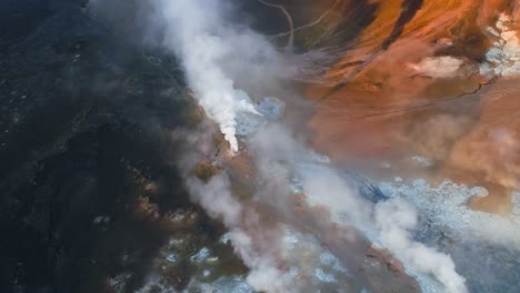 aerial drone footage of hot springs in Greenland or Iceland steaming on mountains with pillars of white smoke coming out of them during a sunny day while footage moves further. Land is orange, black.