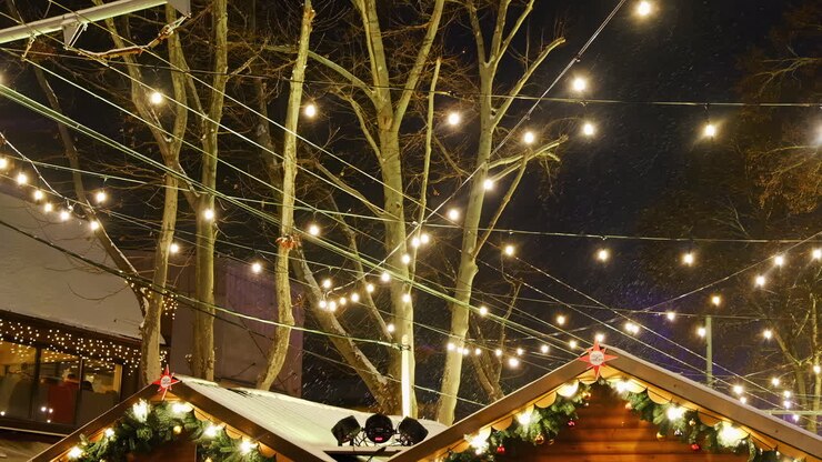 Bright twinkling lights illuminate the holiday market. Stalls adorned with decorations stand among trees, creating a cheerful atmosphere on a winter night