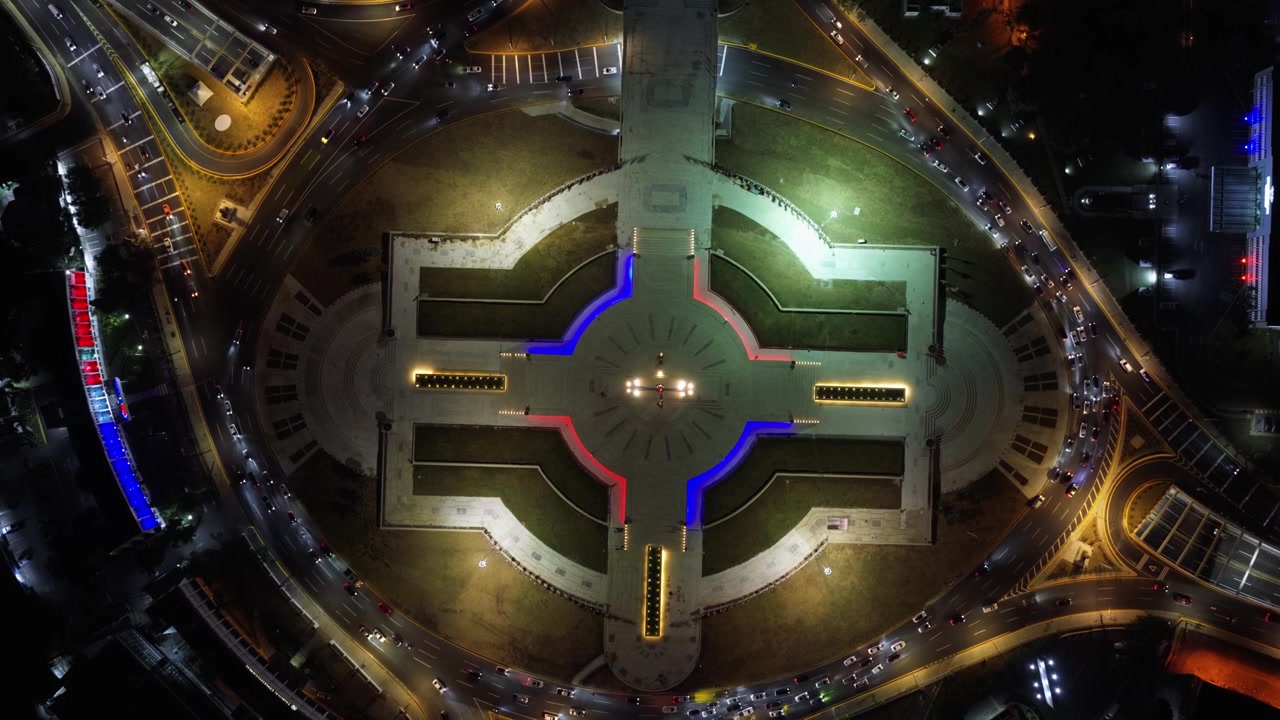 Il monumento alla bandiera illuminato si trova al centro della rotonda di Plaza de la Bandera a Santo Domingo, Repubblica Dominicana; le auto circondano percorsi illuminati di rosso e blu di notte. Vista aerea con drone dall'alto