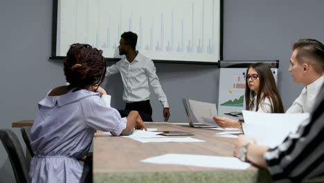 african american coach gives corporate presentation for businesspeople in office, dark skinned businessman presents new business plan on whiteboard, speaking to clients listening speaker at meeting