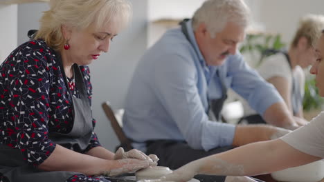 medium shot of middle aged ceramic artist teaching group elderly caucasian woman and senior man how to wedge clay sitting at desk in art studio. people enjoying talking at work