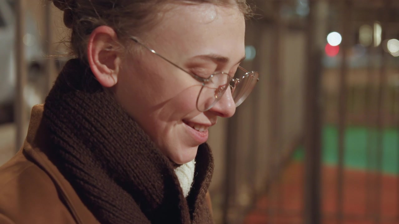 Femme caucasienne en extérieur, la nuit, lisant un carnet dans un microphone. Elle porte un manteau marron et une écharpe épaisse, des lunettes rondes et ses cheveux sont attachés en chignon. Debout près d'une clôture métallique, avec les lumières de la ville en doux bokeh, elle apparaît contemplative.