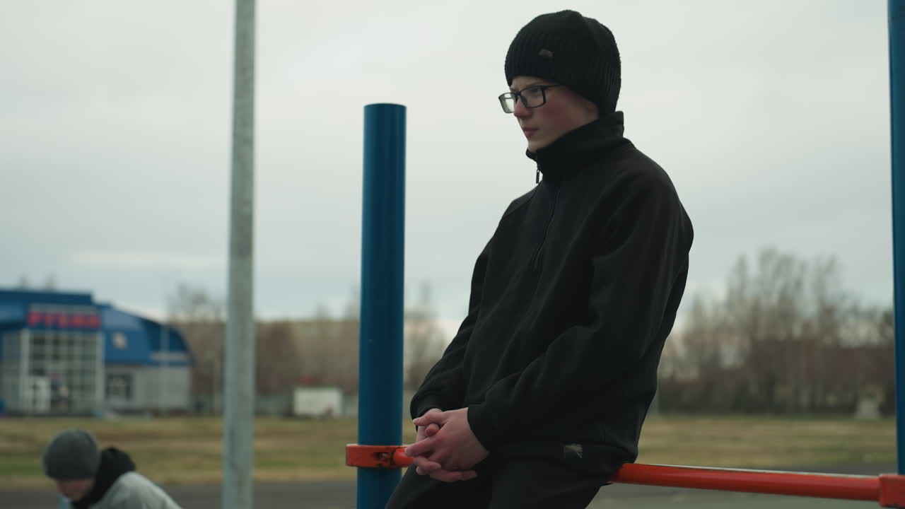 Side view of a young boy wearing a black outfit and glasses, sitting on a red iron bar with his hands clasped together, with a blur view of someone in gray sweater close by