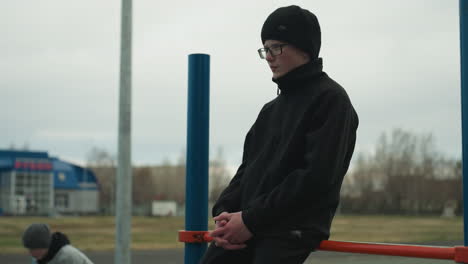 side view of a young boy wearing a black outfit and glasses, sitting on a red iron bar with his hands clasped together, with a blur view of someone in gray sweater close by