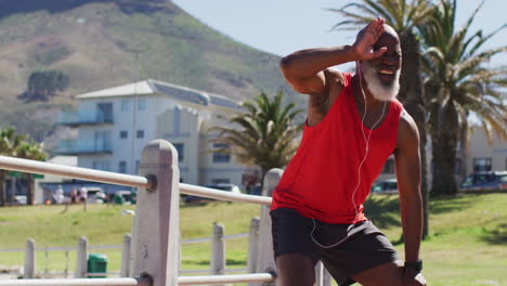 Tired-senior-african-american-man-wearing-headphones-taking-a-break-from-running-on-promenade