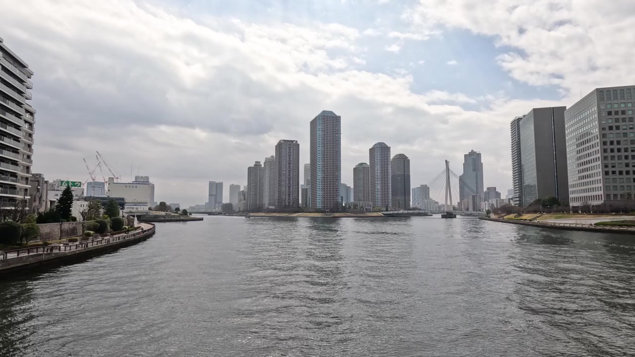 Static wide shot of the Sumida River and Chuo City skyline under cloudy skies.