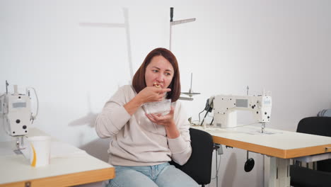 seamstress seated in bright workspace between two knitting machines eating breakfast from clear disposable plate, casual atmosphere with tools and materials around, enjoying peaceful break