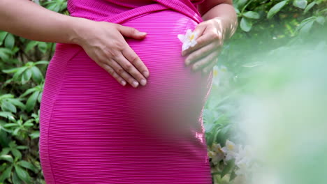 pregnant woman in dress holds hands on belly on natural background of rhododendron at summer day