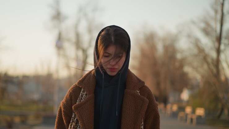 Front shot of a sad girl walking through a lonely park at sunset, wearing a brown coat and a hoodie. melancholic mood with the girl's downcast expression and the serene, empty park setting