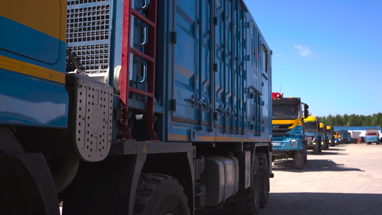 Outdoor yard scene showing blue and yellow heavy-duty truck trailers with corrugated doors, cam locks, hinges, red ladder, perforated step plate, fuel tank, gravel ground, clear sky