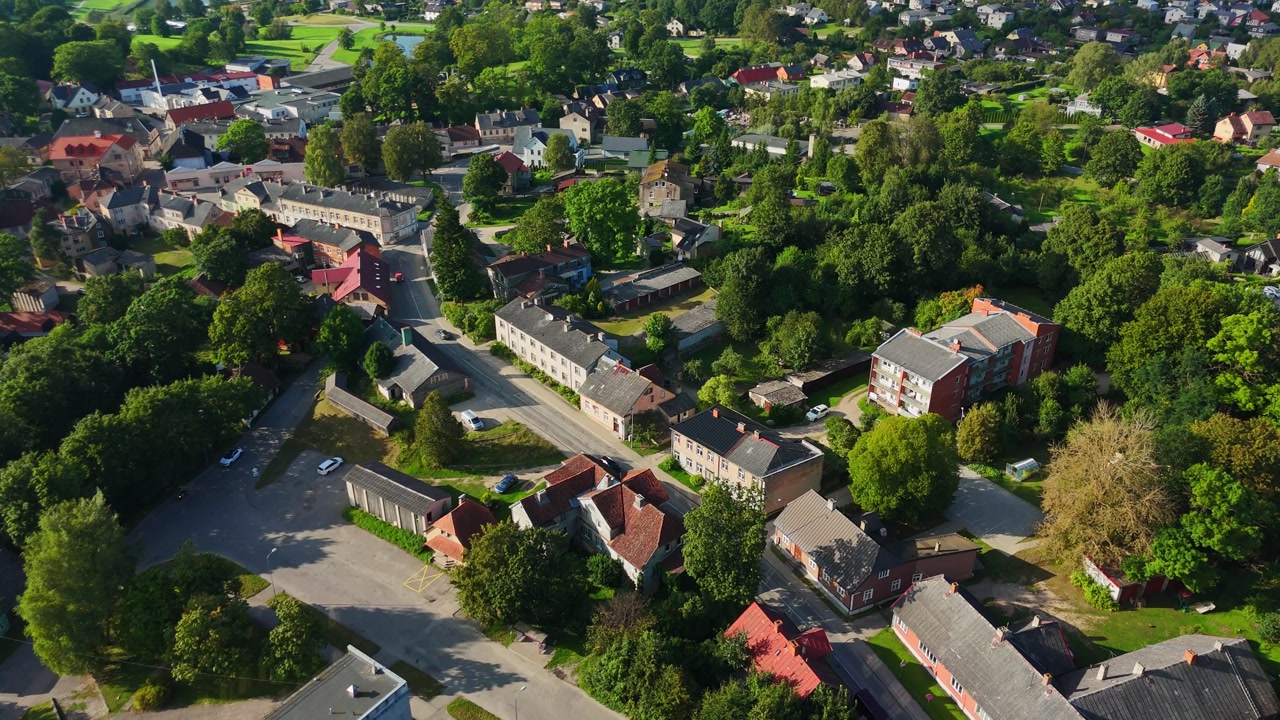 Drone establishing shot revealing Talsi's historic Latvian landscape, showcasing red roofed buildings nestled among verdant greenery under bright summer sunlight