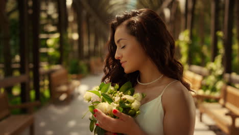 Gorgeous-bride-standing-under-arch.-Beautiful-woman-smelling-flowers-in-garden