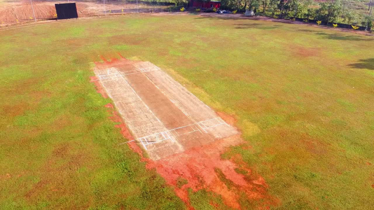 Ripresa aerea con drone di un campo da cricket rurale in una giornata di sole, che mostra il campo rustico, l'outfield erboso e i dintorni di terra rossa. Perfetta per contenuti su sport, viaggi e cultura locale.