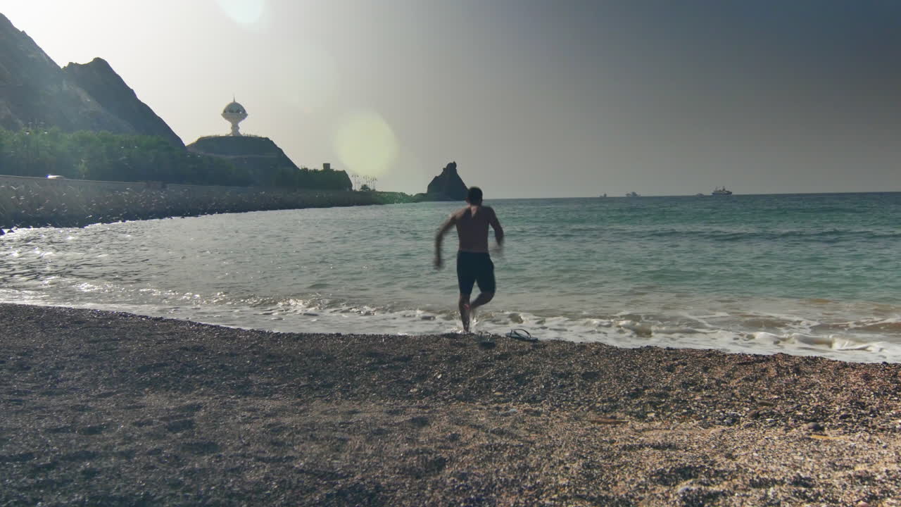 Barefoot man playing by the sea with mountains behind and iconic incense burner in the distance under harsh sunlight