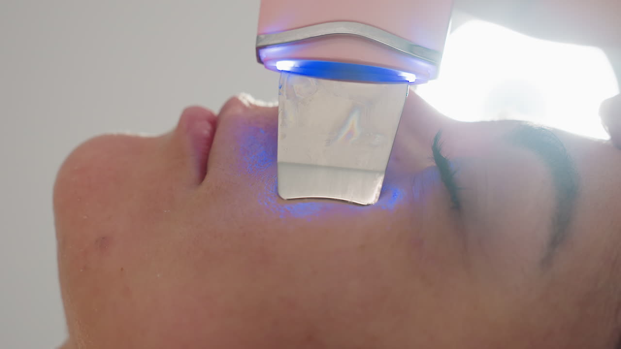 Close up view of technician gliding vibrating ultrasonic tool with blue light across client skin during spa treatment for soothing facial massage and rejuvenation under soft lighting