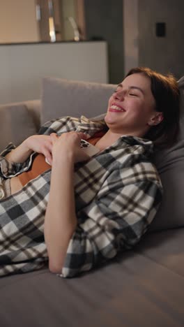 Vertical-video-of-a-happy-and-joyful-brunette-girl-in-a-checkered-shirt-and-an-orange-top-looks-at-the-social-network-using-a-white-smartphone-and-communicates-online-in-a-modern-apartment-lying-on-the-sofa