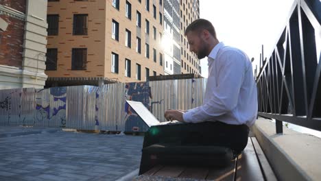 successful entrepreneur works on notebook sitting on bench at city. confident manager typing text on laptop outdoor. young businessman using computer at town. concept of business and finance. close up