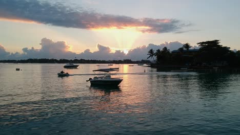 Un-Dron-Disparó-A-Baja-Altura-Moviéndose-Hacia-Un-Pequeño-Bote,-Mientras-Otro-Diminuto-Fuera-De-Borda-Pasaba-Por-La-Escena,-Al-Amanecer-En-Un-Pequeño-Muelle-En-Una-Isla-Paradisíaca