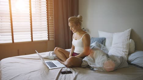 mujer encantadora en pijama sentada en la cama cerca de la computadora portátil y video chateando con sus amigos