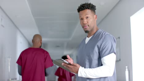 portrait of african american male doctor wearing scrubs, using tablet in corridor, slow motion