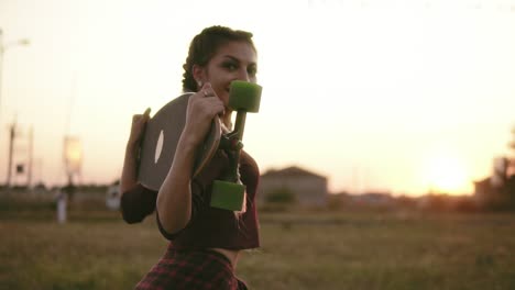 vista trasera de una mujer con camisa a cuadros y camiseta sin mangas sosteniendo longboard y caminando durante la puesta de sol en verano