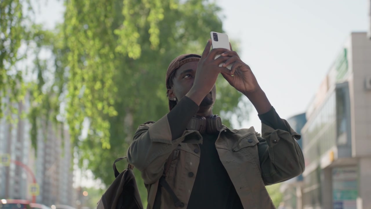 Close up of man holding smartphone in urban street setting with green trees and buildings. Man wears brown cap jacket carrying backpack interacts steadily with device. Soft daylight