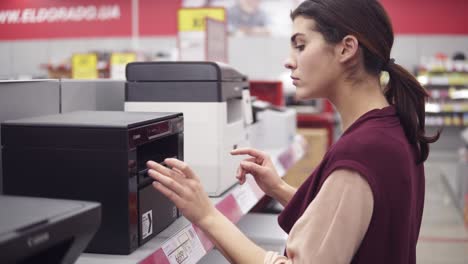 female costumer choosing new printer in electronics department in appliance store. examining it carefuly. looking for household electronics.