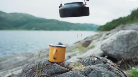 pouring hot coffee from portable coffee pot into mug on rock by the lake shore