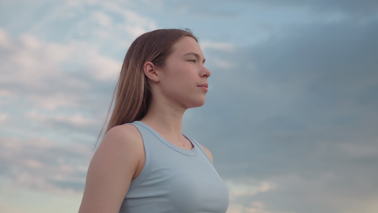 elegant young woman adjusting long hair under cloudy sky, eyes closed in calm expression, wind tousling strands, pastel clouds framing profile, soft light illuminating skin against backdrop