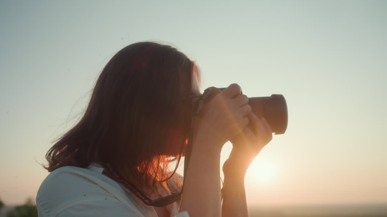 Close up of woman taking photo with camera during golden hour, eyes focused through viewfinder as warm sun glows around hands, blurred tree visible in distant background