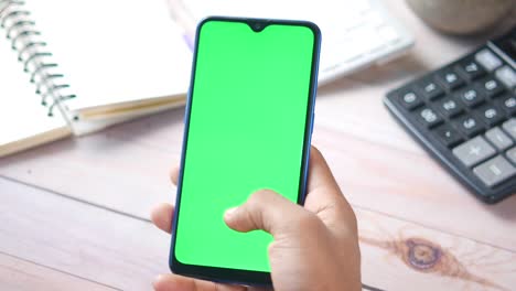 person holding a smartphone with a green screen on a wooden desk