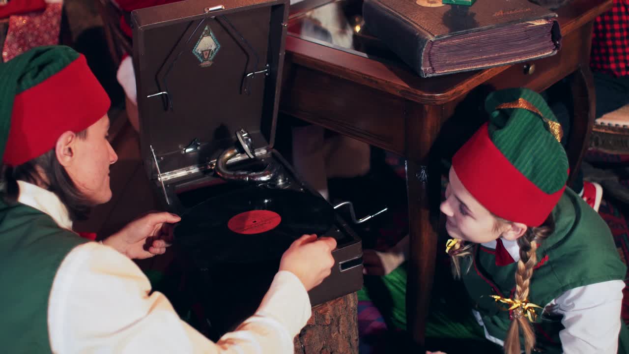 An elf girl in a green suit gives a vinyl record to an elf. Dwarves are listening to an old gramophone