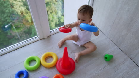 little caucasian boy sits on the floor in the balcony. active toddler plays with toy pyramid. top view.