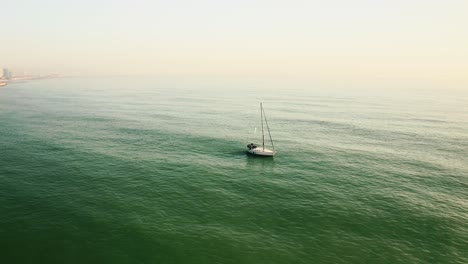 AERIAL:-Small-sailboat-sails-slowly-off-the-coast-of-barcelona,-spain