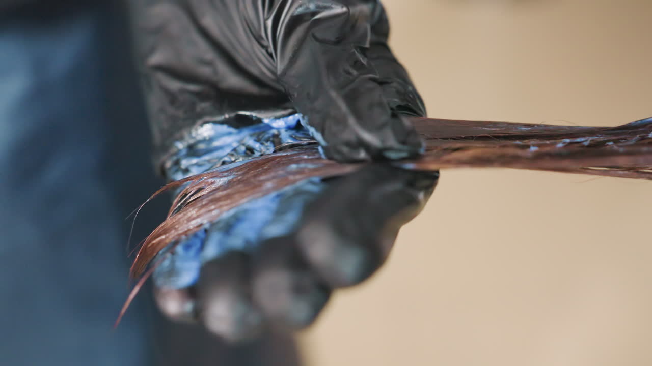 Mano con guanto che dipinge una ciocca blu con un pennello, primo piano dello stylist che applica il pigmento sulle punte, tratti delicati, presa del guanto in lattice, capelli castano lucido, tecnica lenta per una saturazione uniforme in un salone moderno