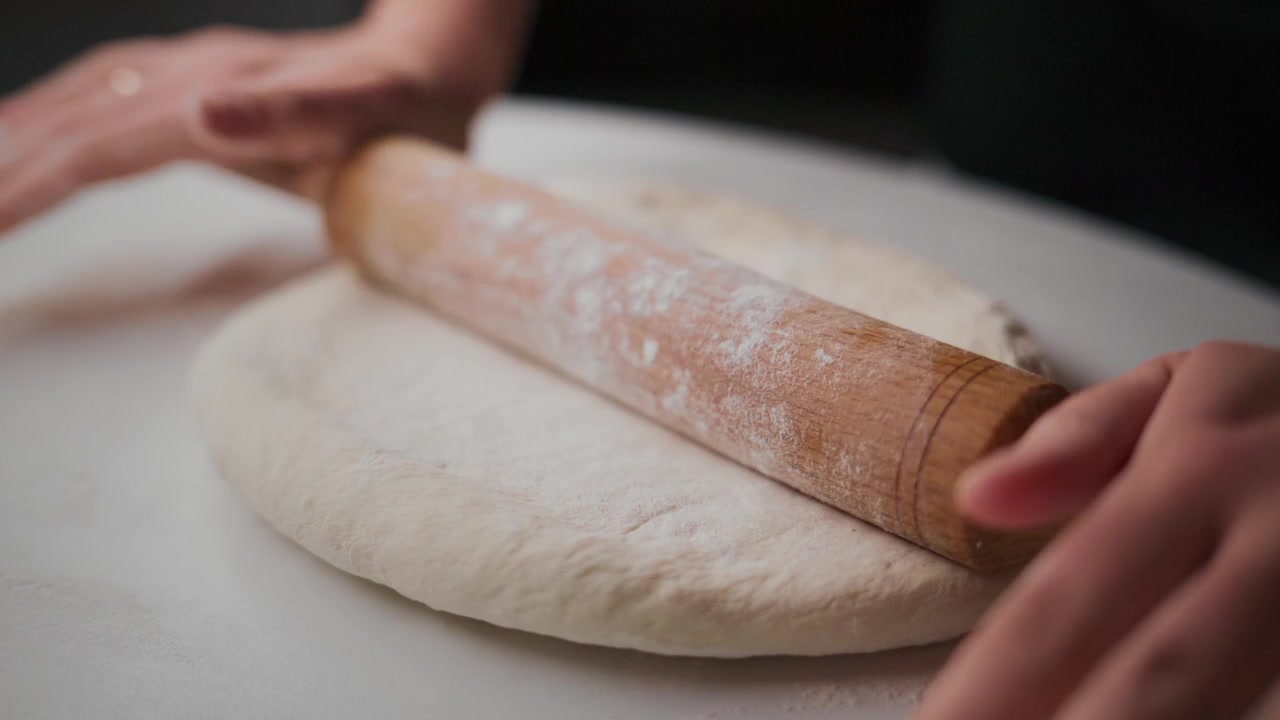 Mani stendono l'impasto su una superficie bianca. Il mattarello appiattisce lentamente l'impasto. Spolverare di farina impedisce all'impasto di attaccarsi. Un primo piano mostra la consistenza dell'impasto. Ideale per cibo, panetteria, cucina o video di ricette.