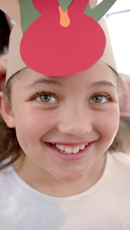 vertical video of happy biracial girl with paper band on head standing and smiling in sunny kitchen