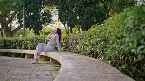 cheerful woman enjoy phone conversation sitting on park bench at green foliage