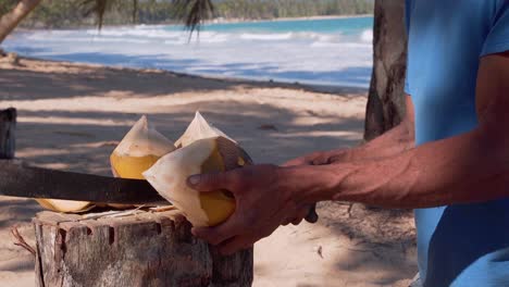 close-up shot in slow motion of man cutting coconut from water, with turquoise blue beach background view and waves hitting the sand in a natural environment