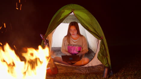 working-remotely-concept-,-young-asiatic-woman-work-at-laptop-form-her-tent-in-front-of-bonfire-while-camping-outdoor