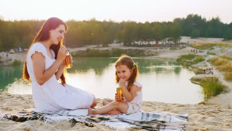 Mama-Mit-Tochter-Drei-Jahre-Entspannt-Am-Strand-Am-See-Trinken-Saft-Aus-Flaschen-Mit-Schlauch