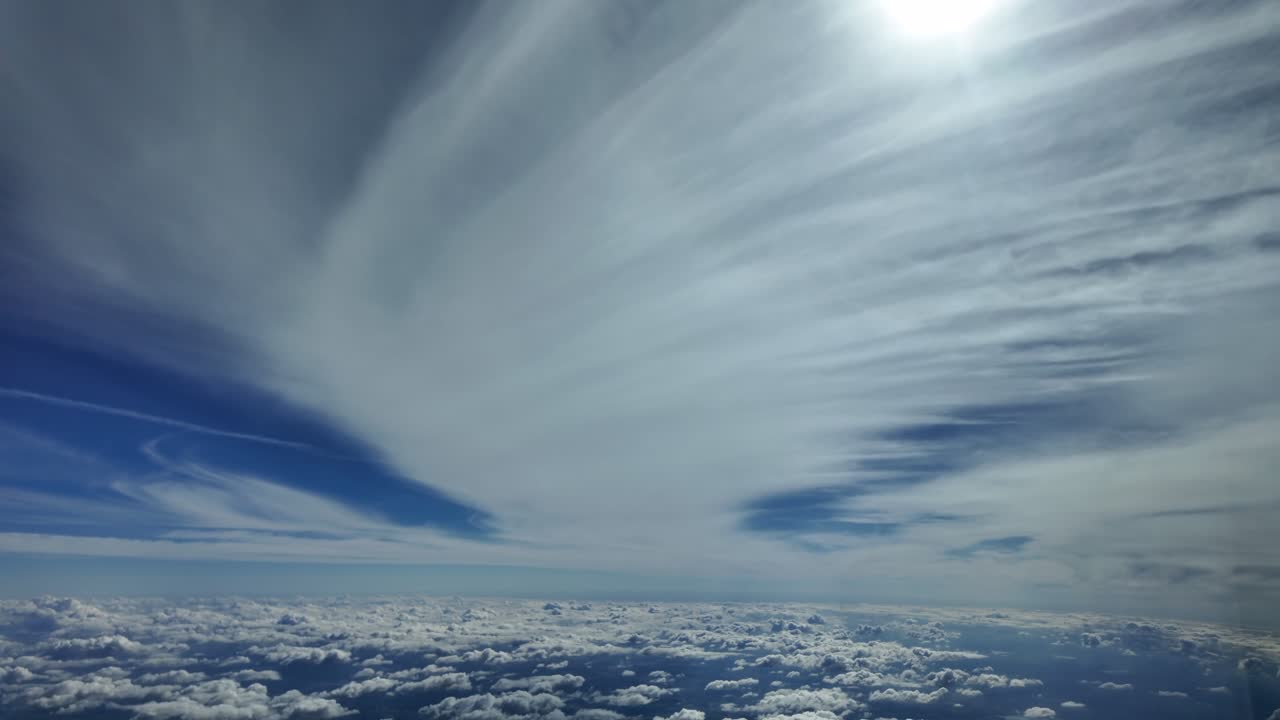 An aerial cockpit view through the pilot’s eyes flying at very high altitude under a layer of unusually shaped cirrus clouds, with a radiant sun shining above in a blue sky.