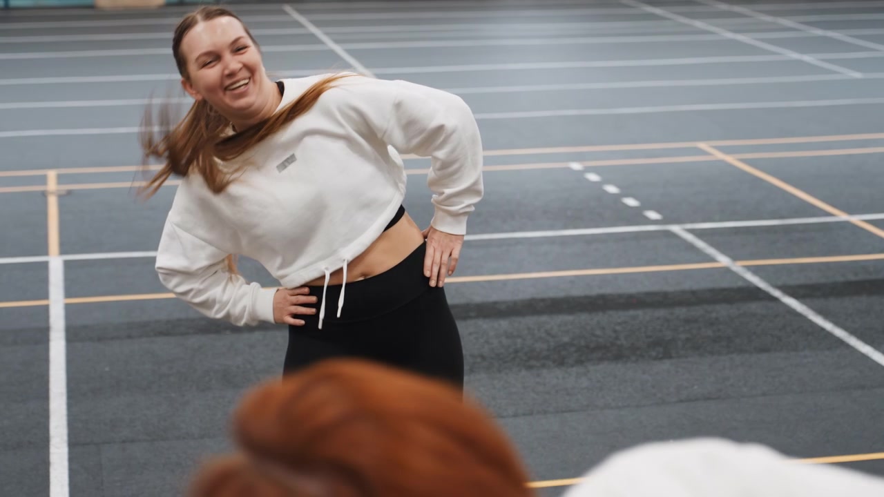 Atleta sorridente si prepara con rotazione del busto. Esercizio di mobilità pre-partita su campo indoor con scala visibile. Calore spensierato mentre l'atleta si allunga da solo prima di una partita di badminton su superficie indoor.