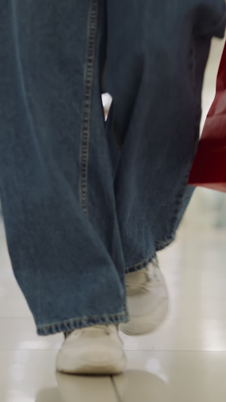Woman legs in loose jeans walk along trade mall hallway closeup slow motion. Lady shopper with purchases in paper shopping bags in business center